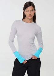 A model wearing a light gray long-sleeve crewneck top with bright turquoise sleeve cuffs, paired with black pants, against a plain white studio background.