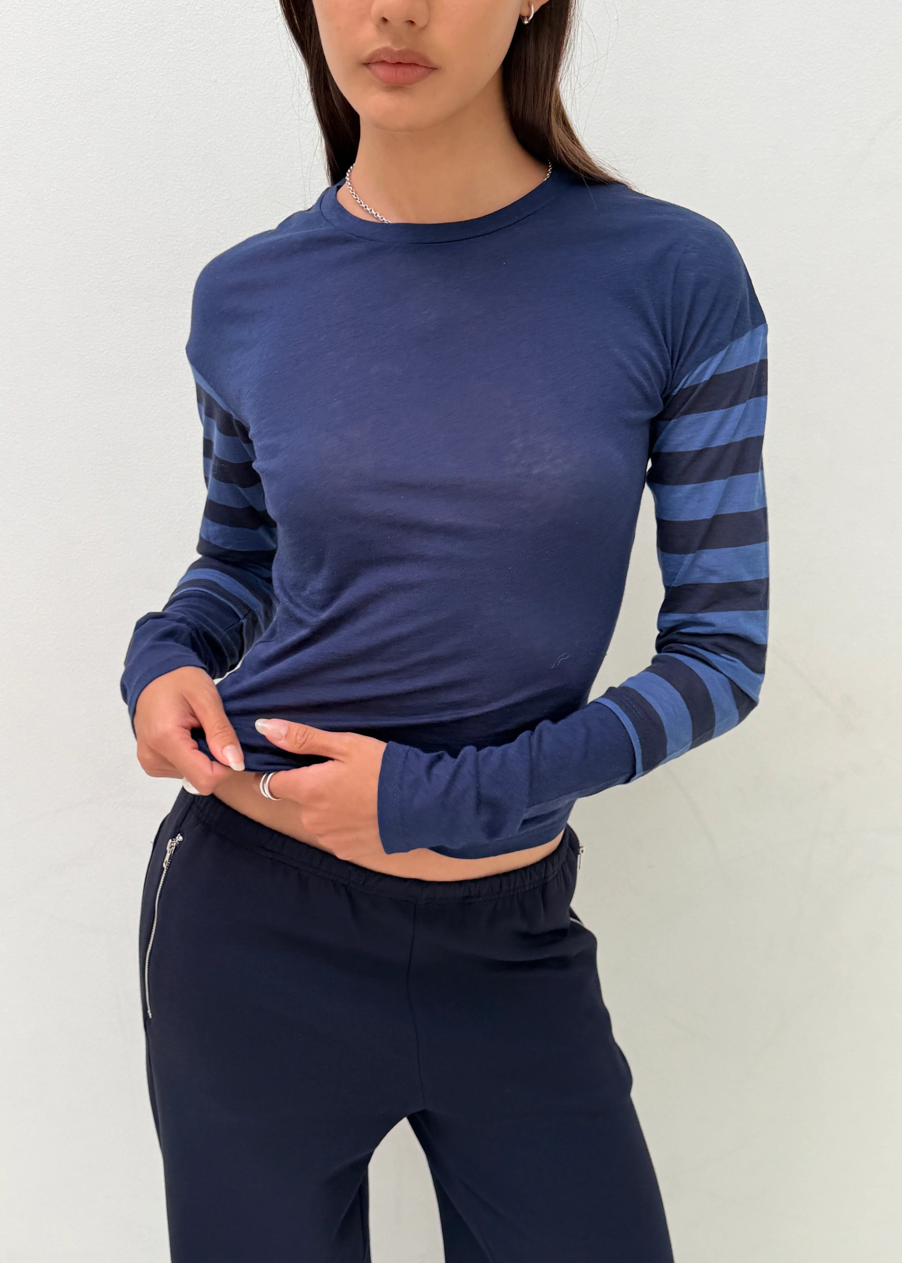 A woman wearing a navy blue long-sleeve crop top with dark blue horizontal striped sleeves, paired with matching navy pants, against a plain off-white background.