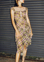 A woman wearing a beige and brown plaid midi dress with a tie-neck detail, styled with black platform sandals, standing in front of a dark corrugated metal shutter.
