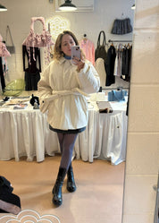 Person taking a mirror selfie wearing a white coat in a clothing store.