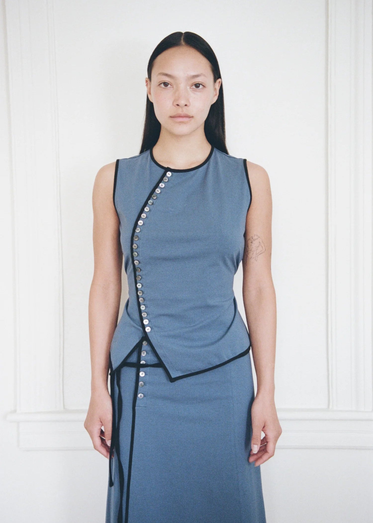 A model stands against a white paneled wall wearing a sleeveless slate blue wrap-style top and matching skirt with black trim and silver button accents.