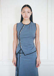 A model stands against a white paneled wall wearing a sleeveless slate blue wrap-style top and matching skirt with black trim and silver button accents.