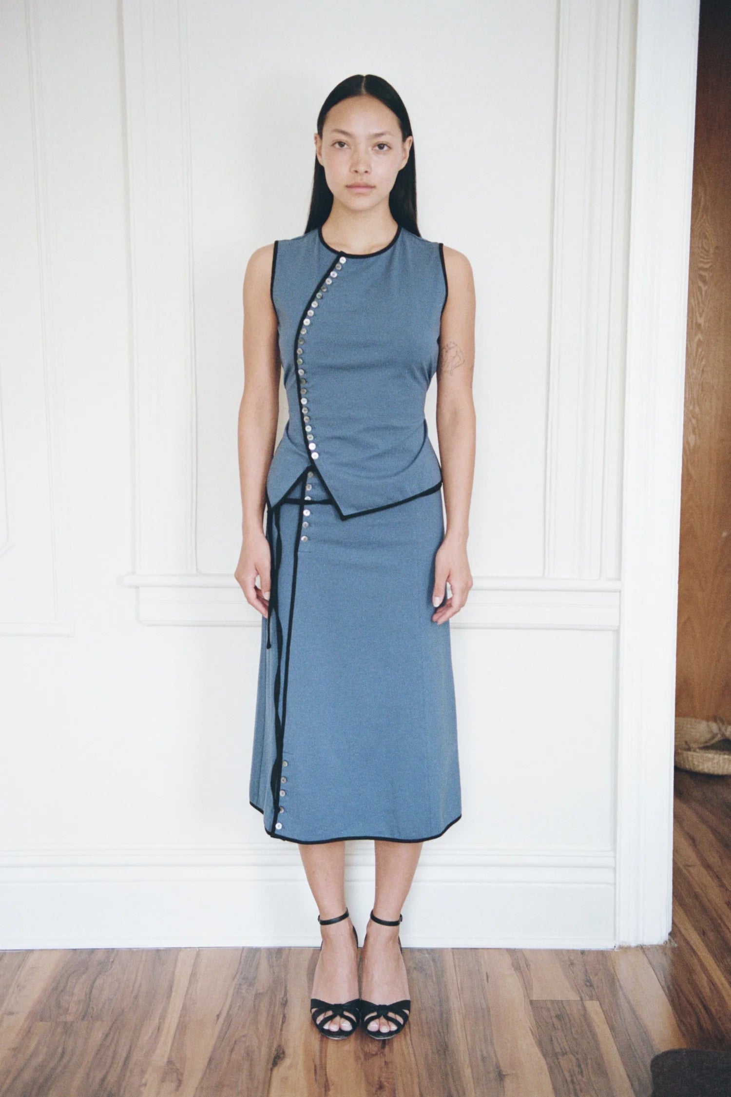 A model stands in a room with white paneled walls and a wooden floor, wearing a sleeveless slate blue dress with black trim and silver-tone decorative buttons down the front.