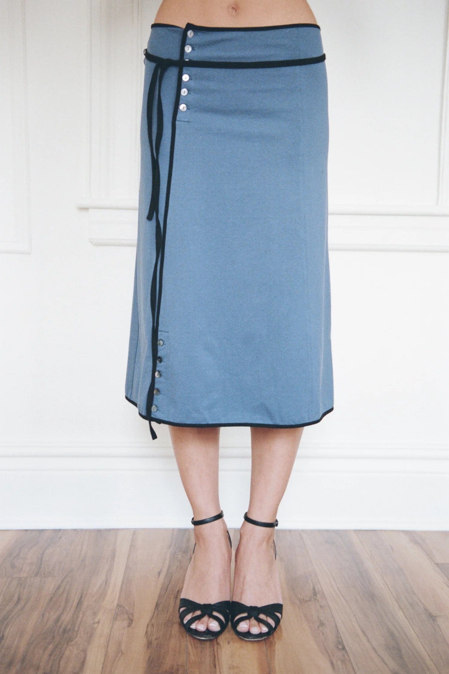 A person wearing a dusty blue wrap-style midi skirt with black trim, side buttons, and a black tie detail, paired with black strappy heels, standing on a wooden floor against a white paneled wall.