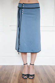 A person wearing a dusty blue wrap-style midi skirt with black trim, side buttons, and a black tie detail, paired with black strappy heels, standing on a wooden floor against a white paneled wall.